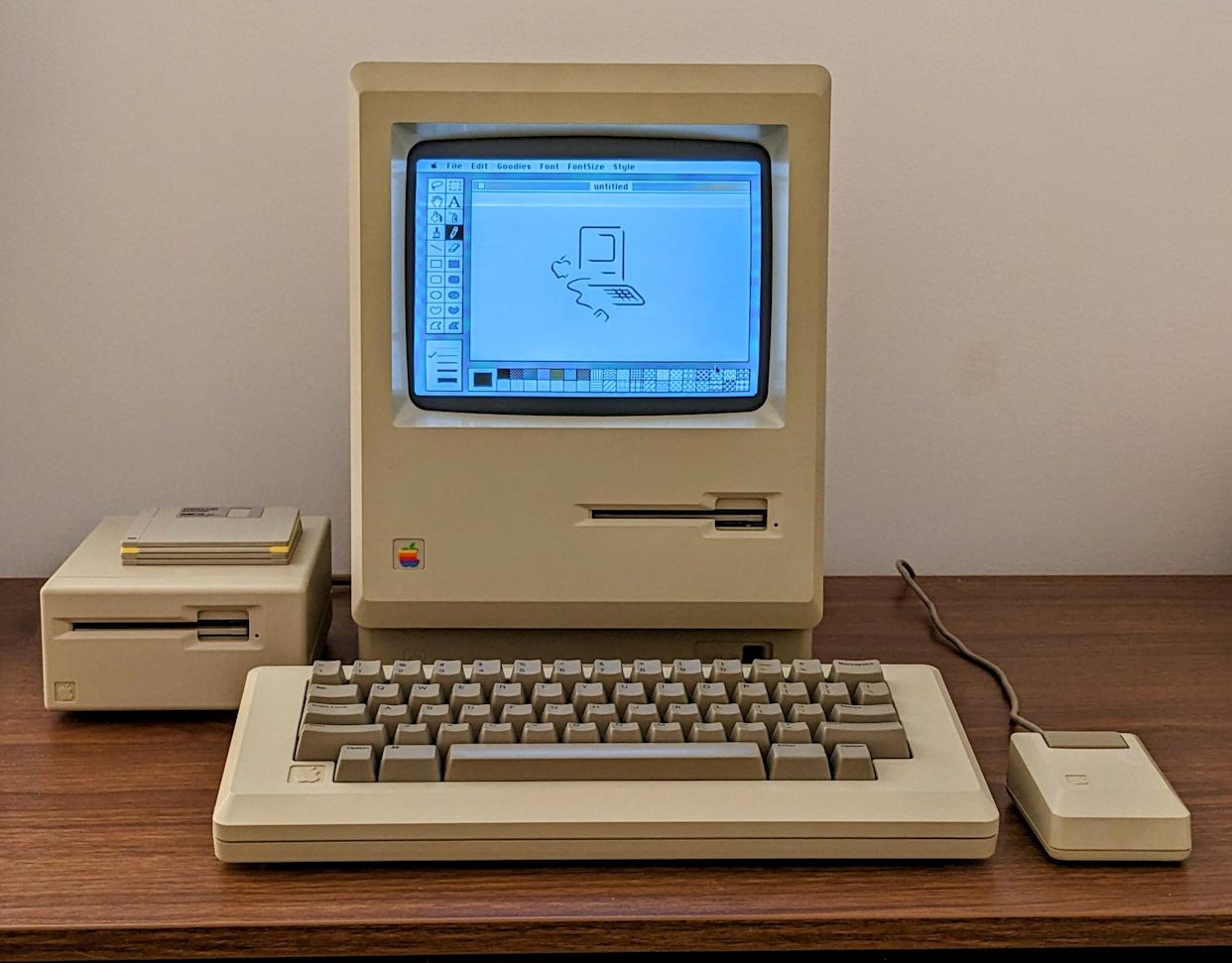An early Apple Macintosh computer with a beige monitor, keyboard, and mouse, sits on a wooden desk. The screen displays a simple black-and-white drawing program with a sketch of a computer. A disk drive is beside the monitor.