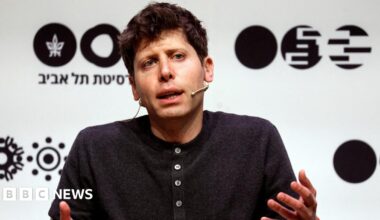 OpenAI leader Sam Altman sitting on a stage, speaking and gesturing with his hands while wearing a dark grey henley sweater.
