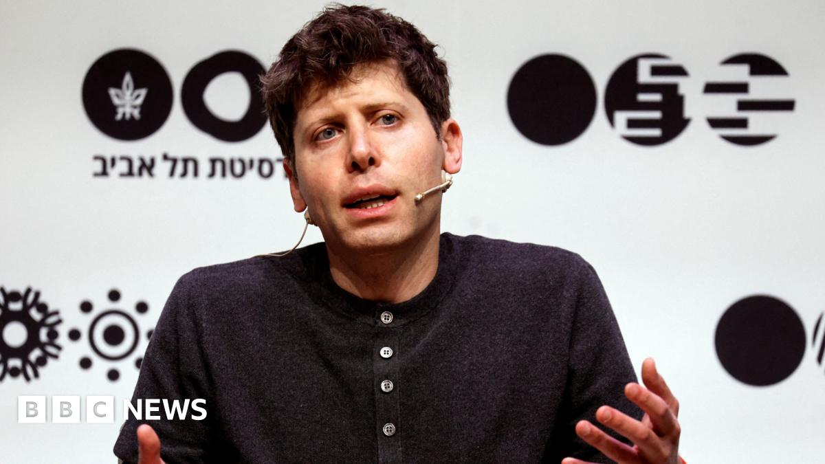 OpenAI leader Sam Altman sitting on a stage, speaking and gesturing with his hands while wearing a dark grey henley sweater.