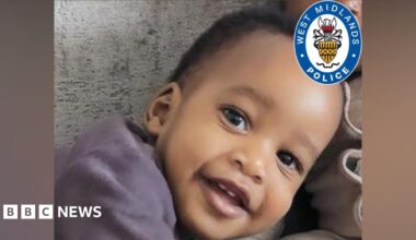 A close up of a baby with short black afro hair. He is leaning to the side and smiling at the camera. In the corner there is a West Midland Police logo