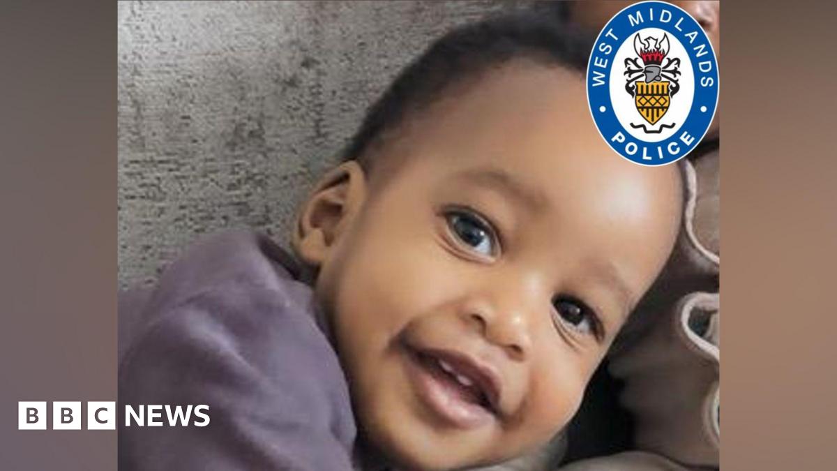 A close up of a baby with short black afro hair. He is leaning to the side and smiling at the camera. In the corner there is a West Midland Police logo