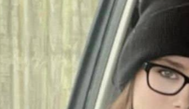 A head and shoulders image of a smiling woman sitting in the front seat of car. She has long brown hair and is wearing black framed spectacles and a black beanie. Her face is turned towards the camera, with the side window behind her.