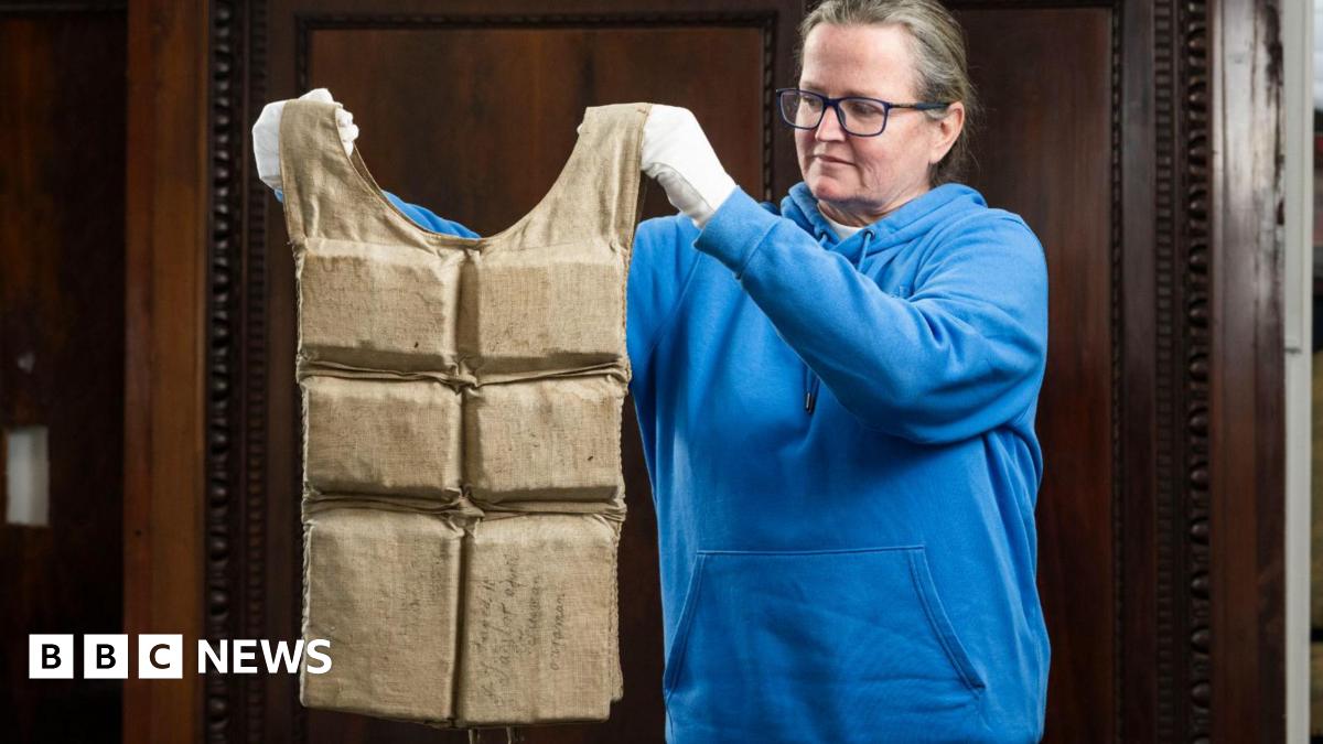 A woman wearing a blue hoodie and white gloves is holding a beige life jacket worn by a Titanic survivor.