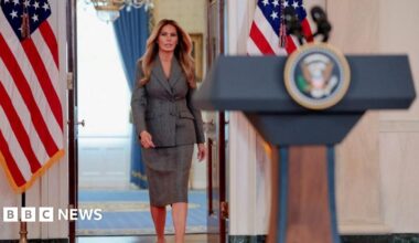 Melania Trump wears a business suit with a skirt and walks toward a podium with the presidential seal at the White House