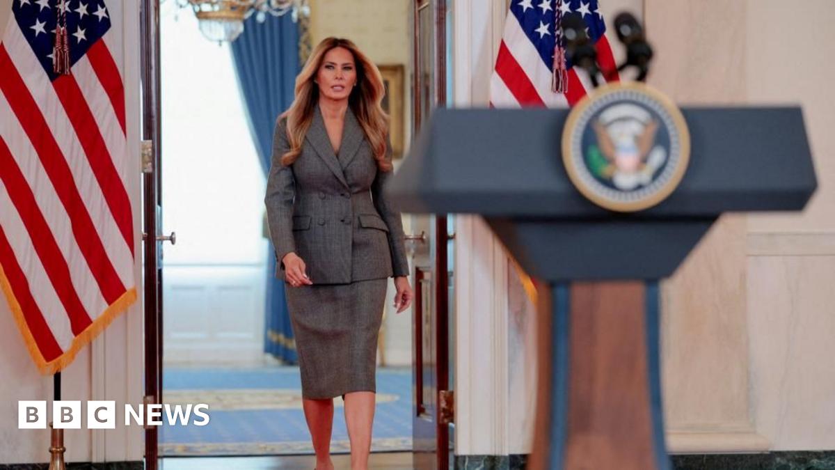 Melania Trump wears a business suit with a skirt and walks toward a podium with the presidential seal at the White House