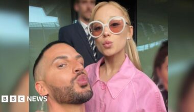 Carl Bembridge poses with the singer Ariana Grande. Carl and Ariana are both pouting and looking at the camera for a selfie. Ariana wears a pink jacket and white rimmed sunglasses.