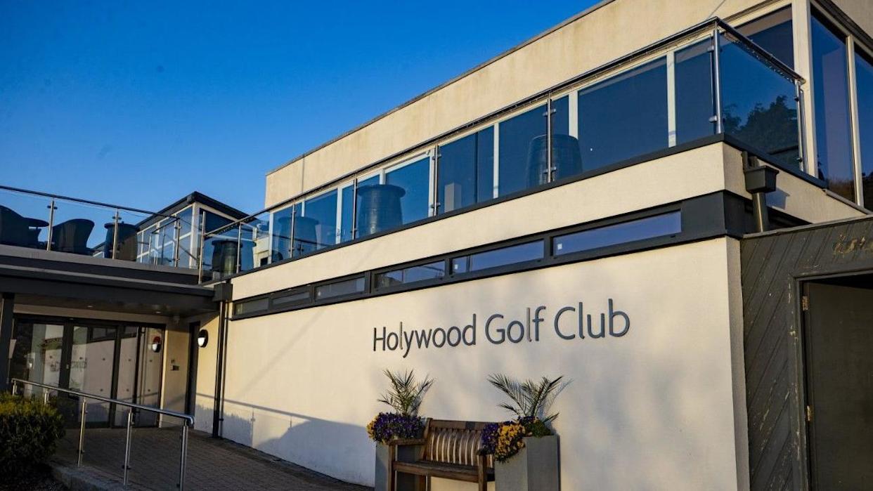 The front of Holywood Golf Club, a beige stone building, with dark lettering saying the words Holywood Golf Club on the front. It has glass boxes on top of the building and there is a brown bench at the front.