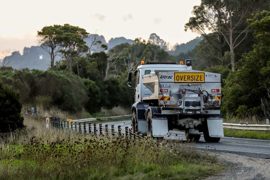 A white truck travels along a rural tree-lined road. It has a yellow sign on the back of its load reading 'OVERSIZE'.