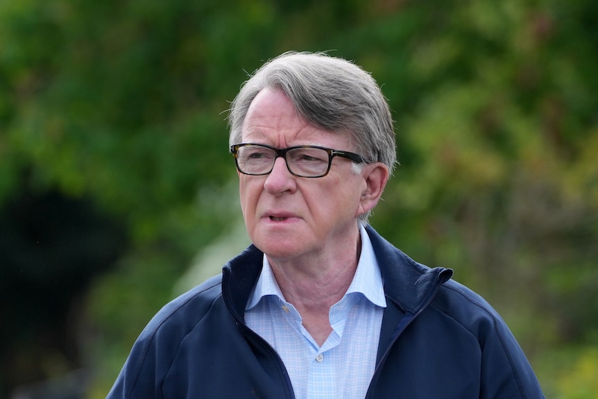 A man with grey hair, black rimmed glasses and a button up shirt seen walking outside with trees behind him.