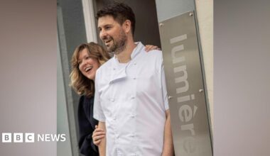 A husband and wife team smile as they stand in the doorway to their restaurant, Lumiere. The woman has mid-length dark blonde hair and is smiling as she stands behind her husband and rests her hands on his shoulder and arm. He has dark hair and stubble, is wearing chefs whites and is smiling.
