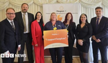 Four women and three men in suits are standing around a woman with a orange sign which reads "I support hospices".