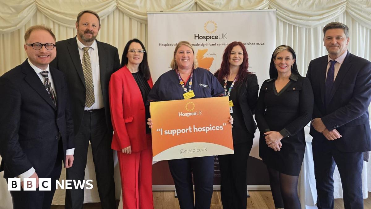 Four women and three men in suits are standing around a woman with a orange sign which reads "I support hospices".