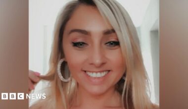 Amber O'Brien smiles as she holds a strand of her blonde hair. She wears large hooped earrings.