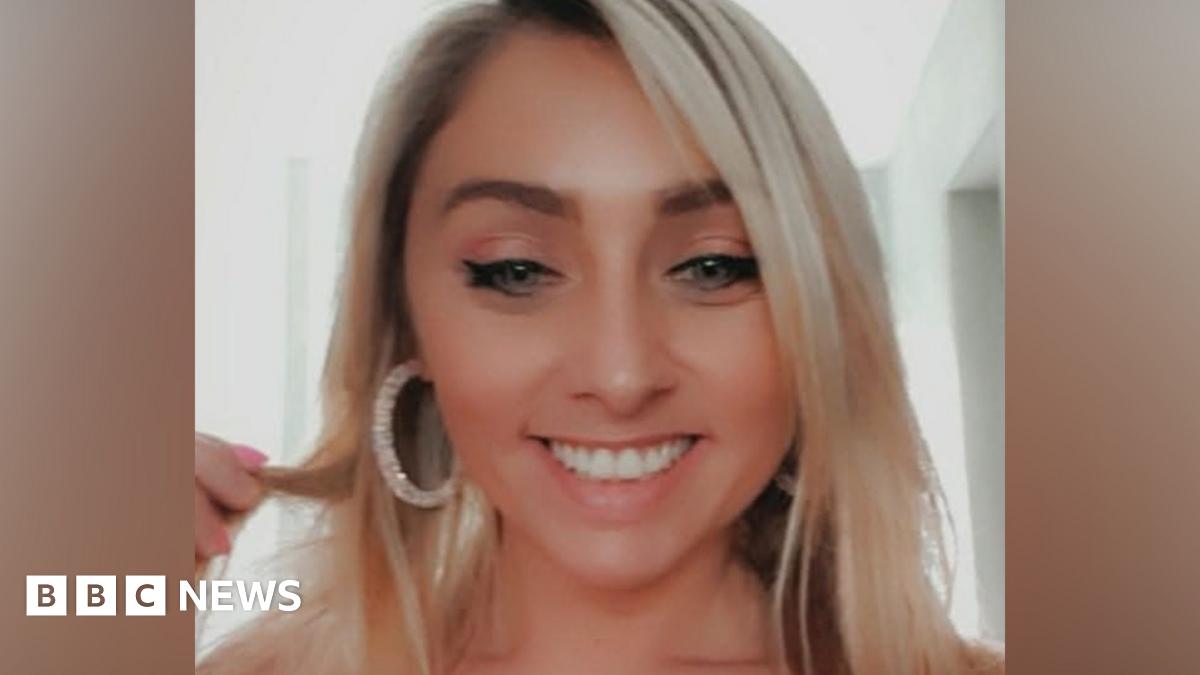 Amber O'Brien smiles as she holds a strand of her blonde hair. She wears large hooped earrings.