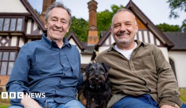 Bob Mortimer and Paul Whitehouse pose for a photo in front of a white house with black dog Ted in the middle.