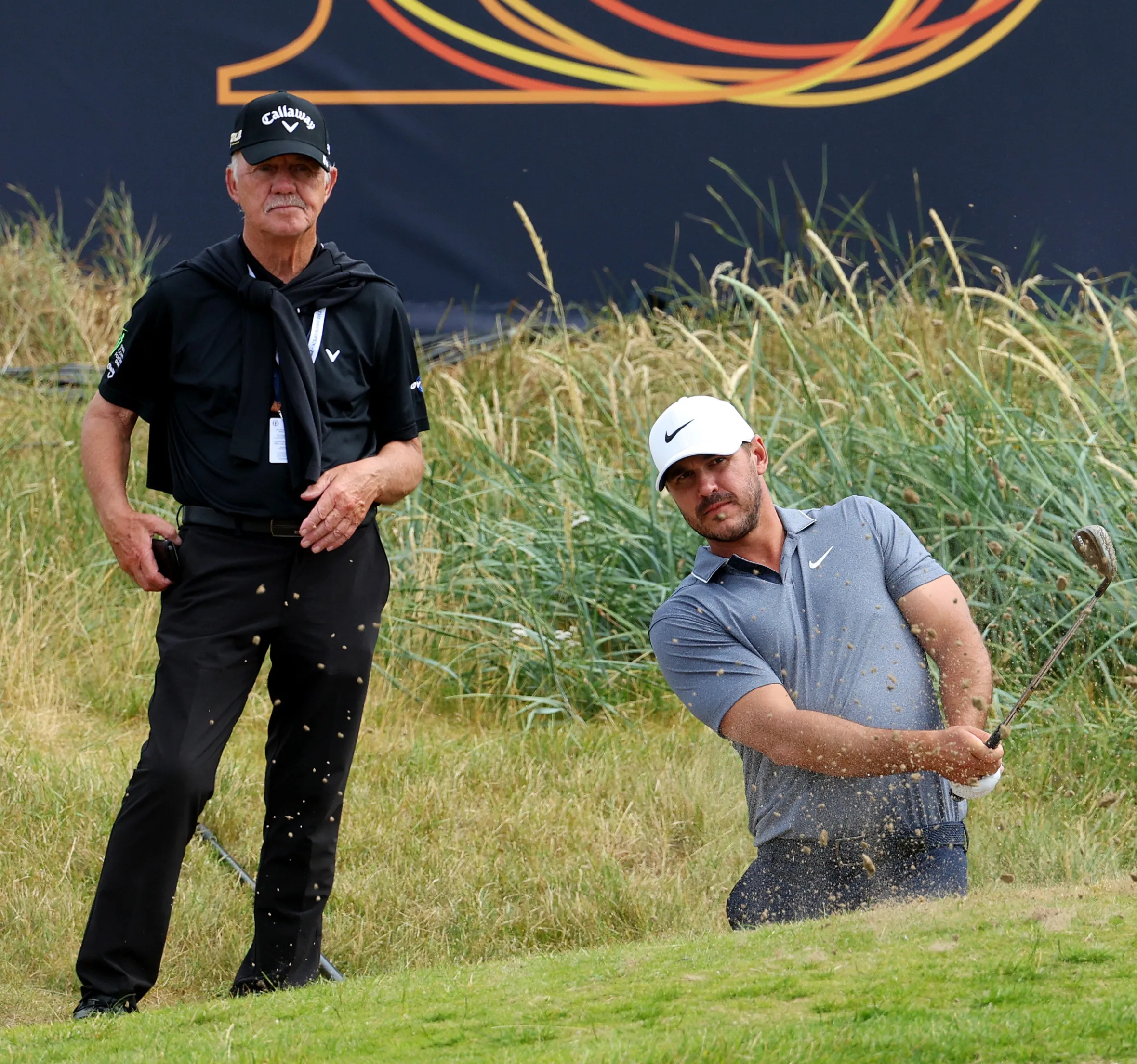 Brooks Koepka chips while Coach Pete Cowen looks on during a practice round at The 150th Open.