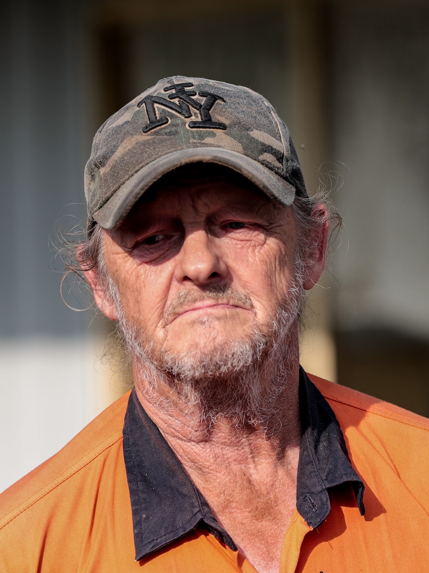 Man wearing a camouflage cap and a orange hi-vis shirt with a blue collar looks ahead, his face is partly shaded. 