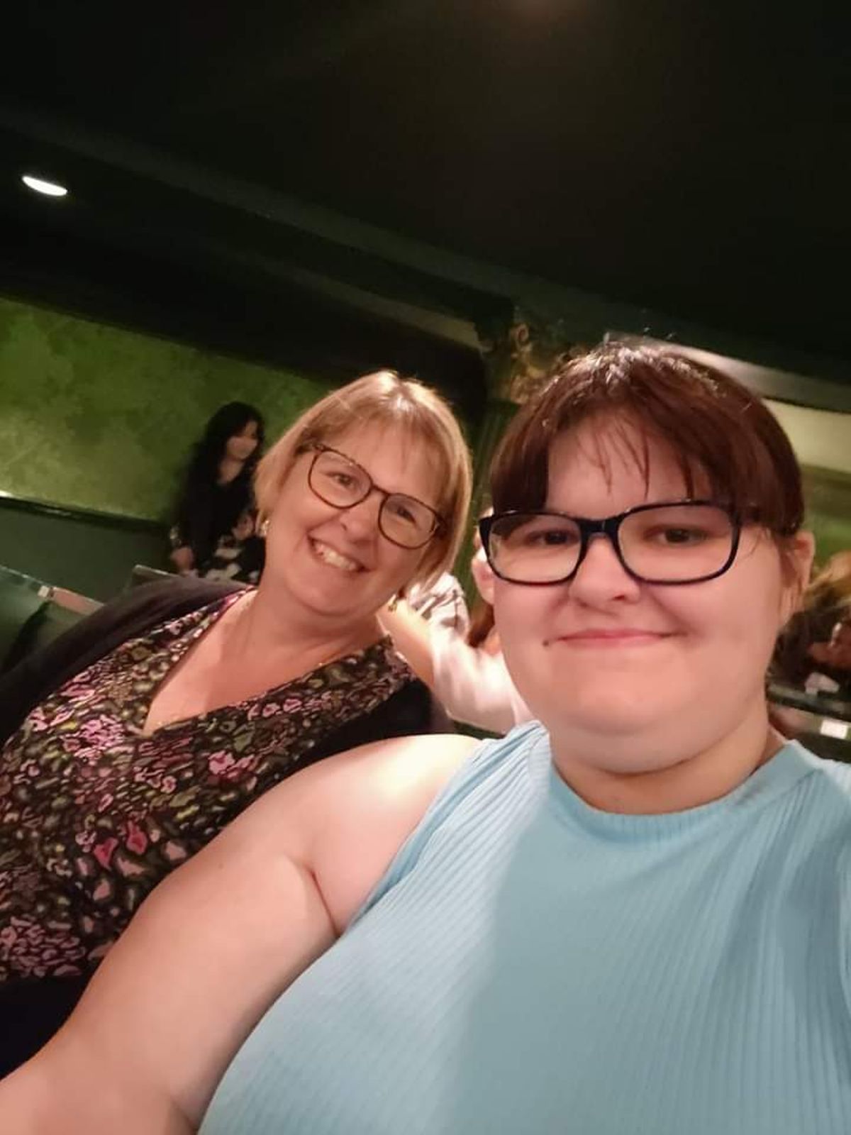 Young woman with glasses in light blue sleeveless top, taking selfie on phone, with woman, also with glasses, in patterned blouse and black cardigan, behind