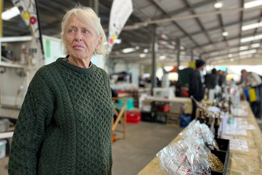 A woman with grey hair stands behind a market stand.