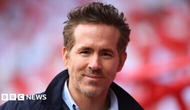 Ryan Reynolds looking directly at the camera, smiling. He has short light brown hair and facial hair. He is wearing a dark navy jacket and a shirt.