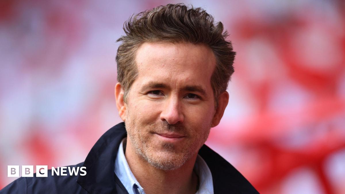 Ryan Reynolds looking directly at the camera, smiling. He has short light brown hair and facial hair. He is wearing a dark navy jacket and a shirt.