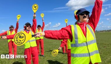 A group of people are dressed in red jumpsuits with yellow high-vis vests holding up yellow signals.