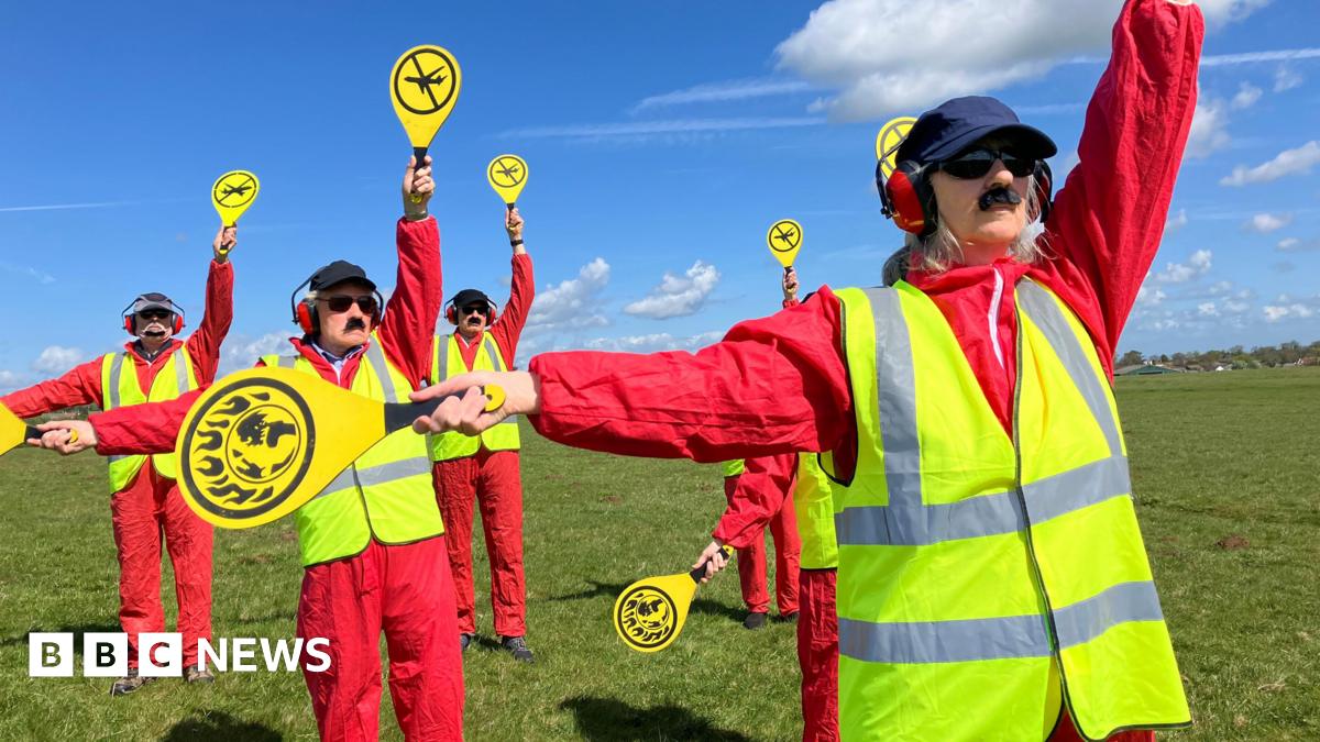 A group of people are dressed in red jumpsuits with yellow high-vis vests holding up yellow signals.