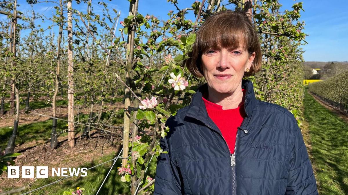 Ali Capper stands on her farm, looking at the camera wearing a dark blue jacket over a red top. She has cropped brown hair.