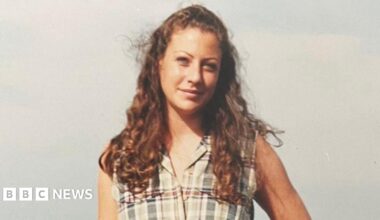 A photo of a young woman with long auburn curly hair wearing a pale checked sleeveless top