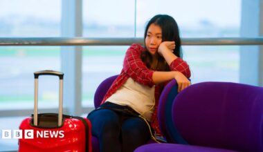 Young woman in red checked shirt rests her chin on her hand. She is sitting in an airport waiting room with a red case next to her