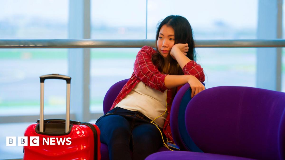 Young woman in red checked shirt rests her chin on her hand. She is sitting in an airport waiting room with a red case next to her