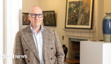 Steve Swallow. He is bald and has blue eyes. He wears glasses with a blue frame, a white shirt and chequered beige and brown jacket. He is leaning on a wall with artworks in the background.