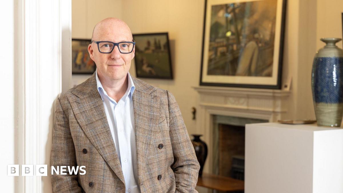 Steve Swallow. He is bald and has blue eyes. He wears glasses with a blue frame, a white shirt and chequered beige and brown jacket. He is leaning on a wall with artworks in the background.