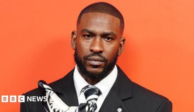 Skepta at the GQ Men of the Year awards. He is wearing a black suit and stood against a bright orange background.