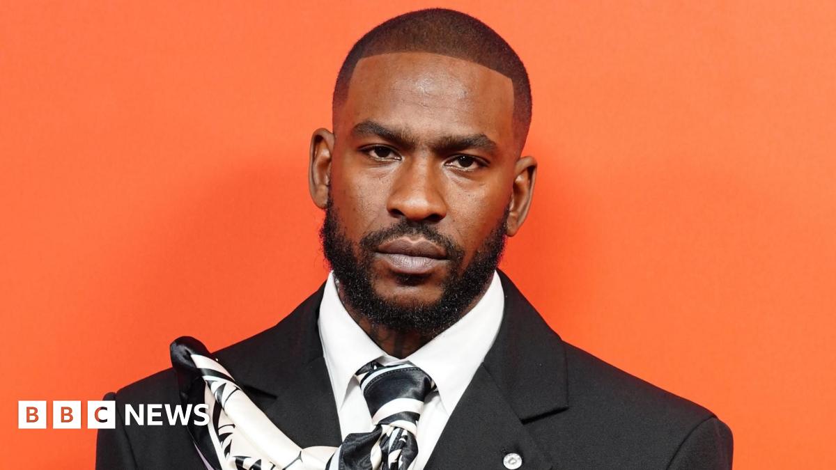 Skepta at the GQ Men of the Year awards. He is wearing a black suit and stood against a bright orange background.