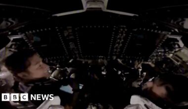 Two astronauts sitting in a dark spacecraft are visible among control systems.