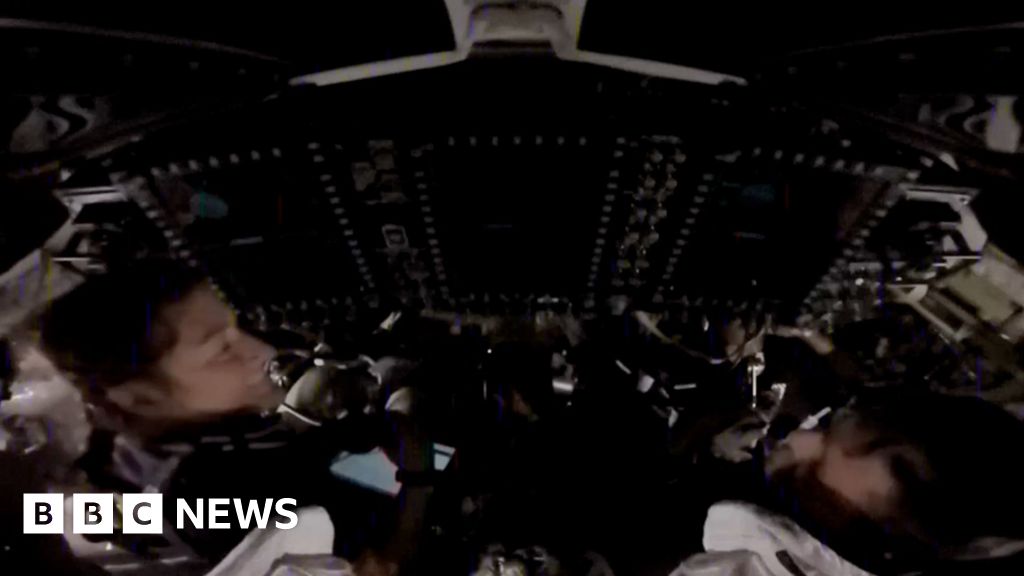 Two astronauts sitting in a dark spacecraft are visible among control systems.