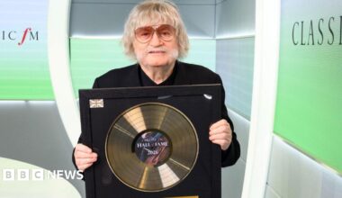 A grey haired man with a big grey moustache - wearing orange shaded sunglasses - holds up his Hall of Fame gold vinyl trophy. He stands in front of a branded screened booth, reading 'Classic FM'.