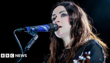 Amy Macdonald performs onstage, singing into a microphone. She has long dark hair and is playing a guitar