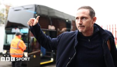 Frank Lampard gives a thumbs up in the direction of a bus outside a stadium.