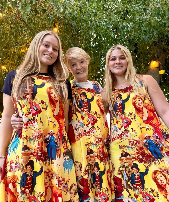 Retired actress and singer Shani Wallis with her two grandchildren, all three wearing matching yellow dresses with illustrations from the musical "Oliver!"