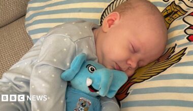 A newborn baby wearing a blue onesie with white stars lies on a blue and white stripy pillow, holding an elephant toy reading "Coventry City"