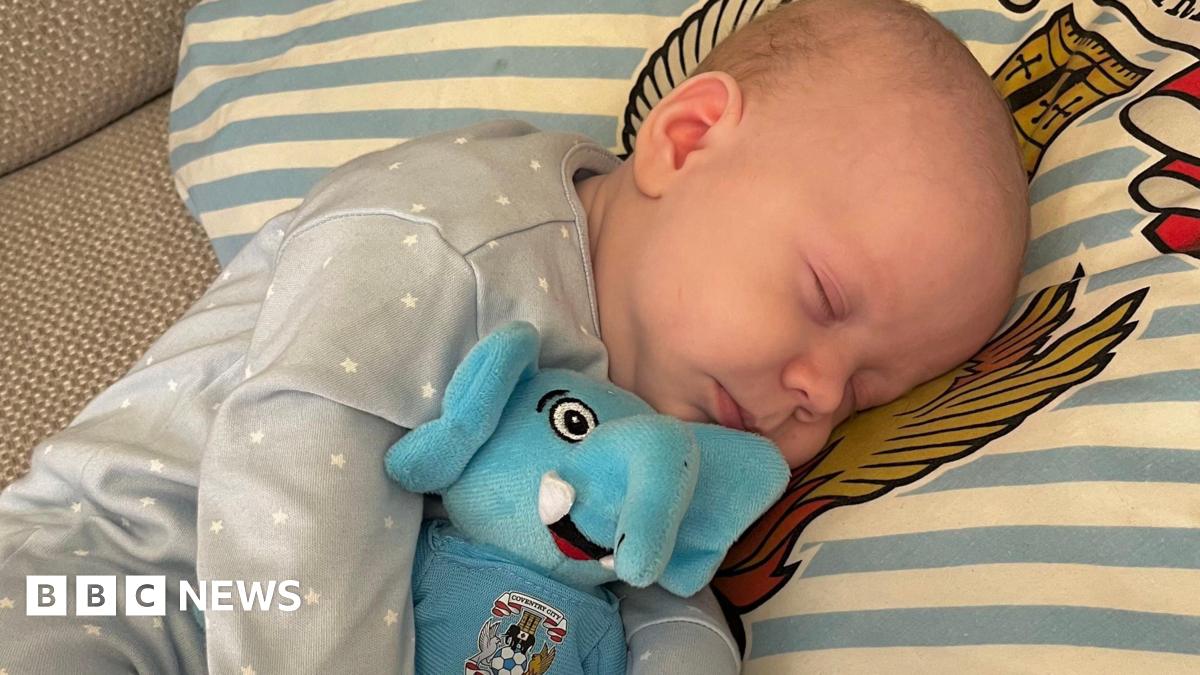 A newborn baby wearing a blue onesie with white stars lies on a blue and white stripy pillow, holding an elephant toy reading "Coventry City"