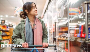 A woman holding a trolley in a supermarket looking to refrigerated shelves.