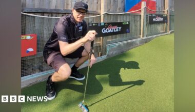 A man in all black squatting by the side of a mini golf course. He is holding a putter.