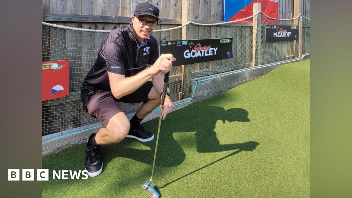 A man in all black squatting by the side of a mini golf course. He is holding a putter.