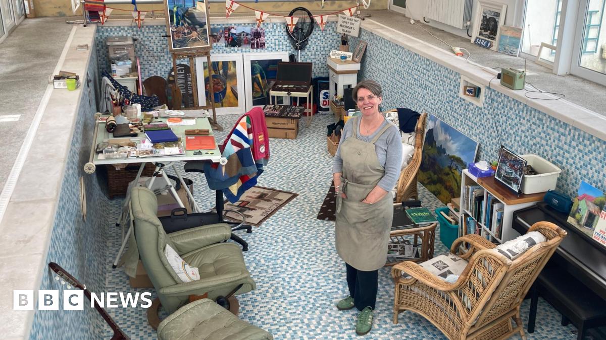 A woman with short grayish brown hair standing in a pool which is filled with various chairs, equipment and paintings. It's arranged so it creates an art studio.