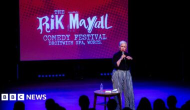 A woman with very short bleach blonde hair and red lipstick is standing on a stage and is speaking into a microphone. She is wearing a black top and black and white striped trousers. Next to her is a small round wooden table with a notebook and water bottle on it. Behind her on a large screen is a red picture with white writing that says "The Rik Mayall comedy festival, Droitwich Spa, Worcs".