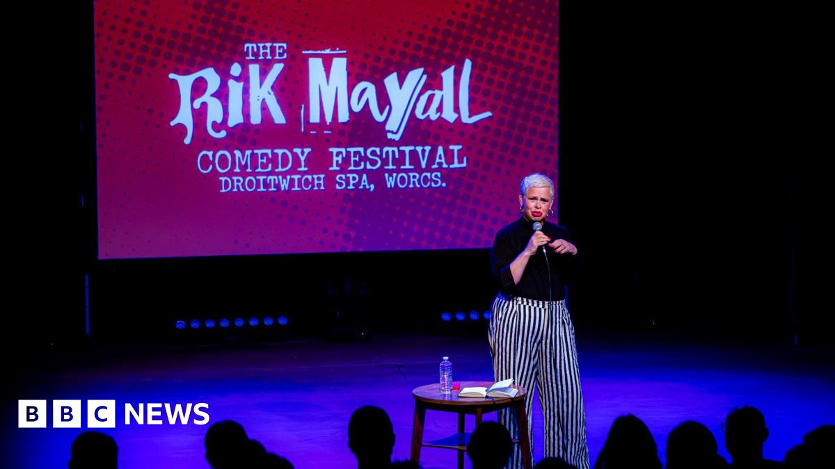A woman with very short bleach blonde hair and red lipstick is standing on a stage and is speaking into a microphone. She is wearing a black top and black and white striped trousers. Next to her is a small round wooden table with a notebook and water bottle on it. Behind her on a large screen is a red picture with white writing that says "The Rik Mayall comedy festival, Droitwich Spa, Worcs".