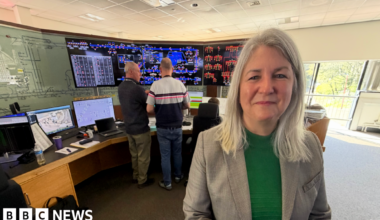 Sarah McManus, who is wearing a green jumper and a grey blazer, stands in front of the soon to be decommissioned system at the control room in South Gosforth. In the background are two controllers who are discussing how the network is currently operating.